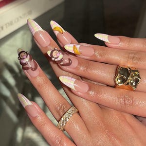 Könnte beinhalten: Lange, mandelförmige Acrylnägel mit hellrosa Grundierung, weißen Spitzen und einem gelb-rosa Karomuster. Einige Nägel zeigen 3D-Amulette von Affen und Bananen, und die Hand ist mit goldenen Ringen geschmückt.
