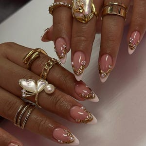 May include: Close-up of multiple gold rings and long, almond-shaped nails. The nails feature a pink base with white tips, adorned with gold embellishments and small pearls. Rings include a butterfly design, bands, and a large pearl ring.
