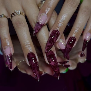 May include: Long, burgundy-colored acrylic nails with a velvet floral design. Some nails have a nude base with gold accents. The nails are long and pointed, with a glossy finish. Gold rings adorn the fingers.