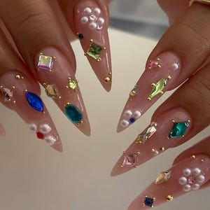 May include: Close-up of long, pointed, manicured nails. Each nail is painted a light pink and adorned with an array of colorful gemstones, pearls, and gold studs. The embellishments include various shapes and sizes, creating a glamorous and detailed design.