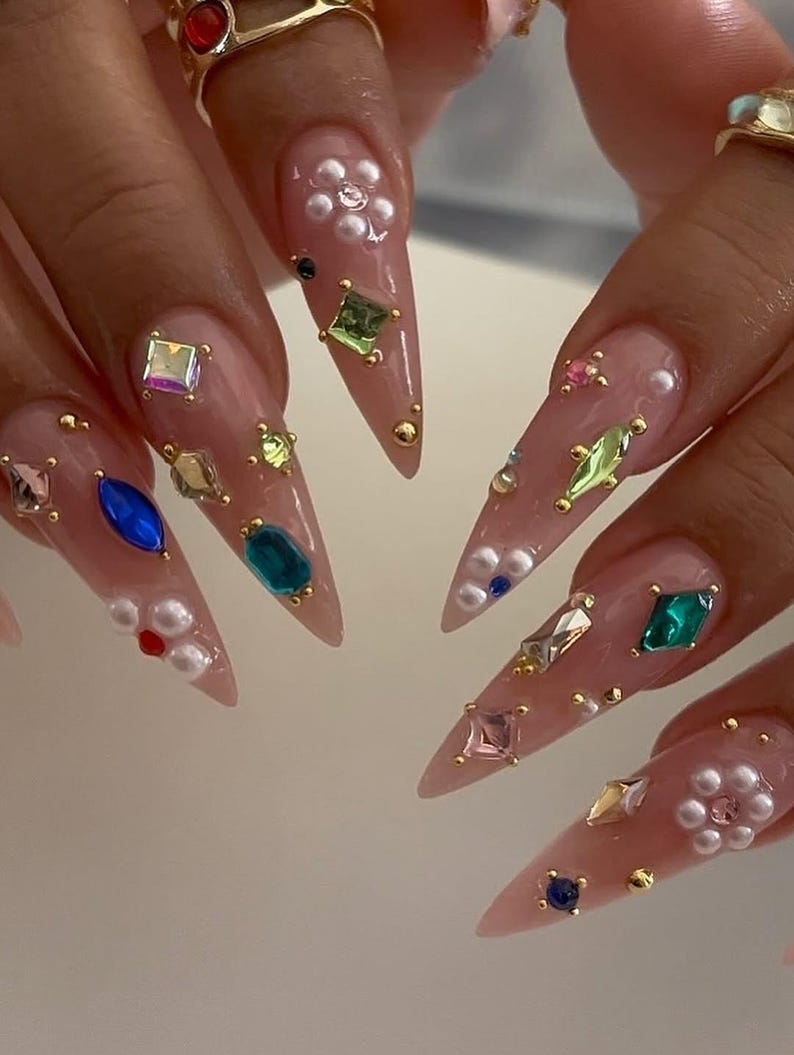 May include: Close-up of long, pointed, manicured nails. Each nail is painted a light pink and adorned with an array of colorful gemstones, pearls, and gold studs. The embellishments include various shapes and sizes, creating a glamorous and detailed design.