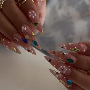May include: Close-up of long, pointed acrylic nails adorned with colorful gemstones, pearls, and gold accents. The nails are a light pink color, with various shapes and sizes of gems. Rings with colorful stones are also visible.