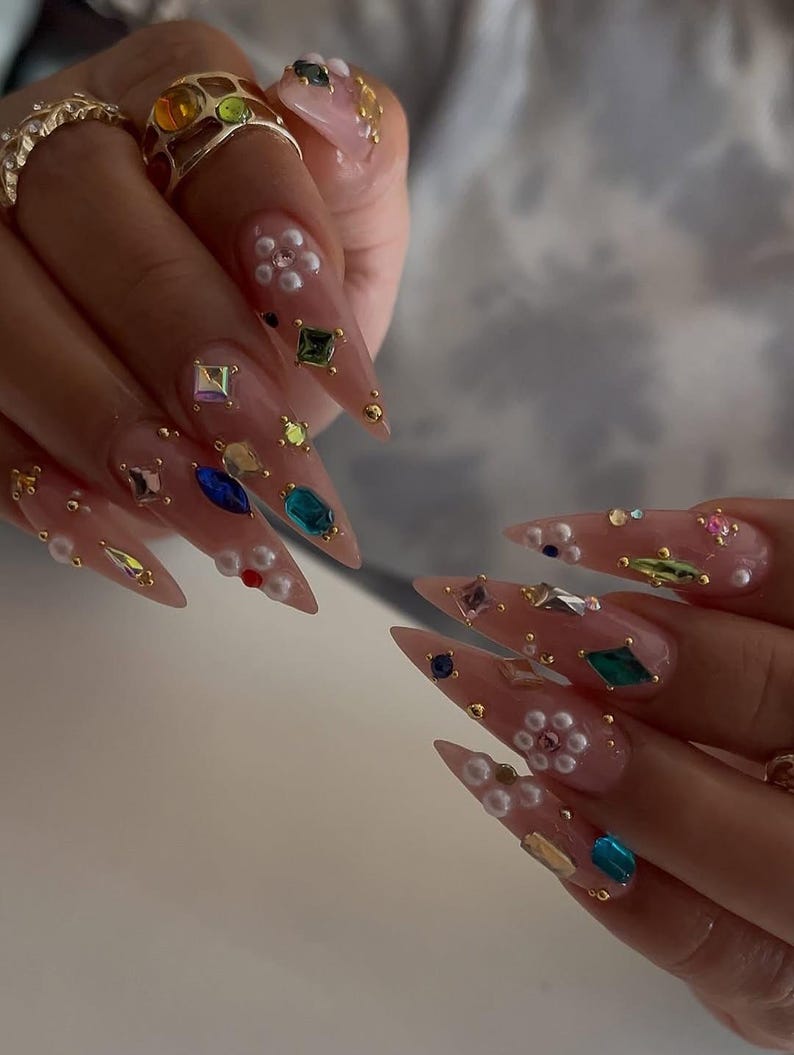 May include: Close-up of long, pointed acrylic nails adorned with colorful gemstones, pearls, and gold accents. The nails are a light pink color, with various shapes and sizes of gems. Rings with colorful stones are also visible.