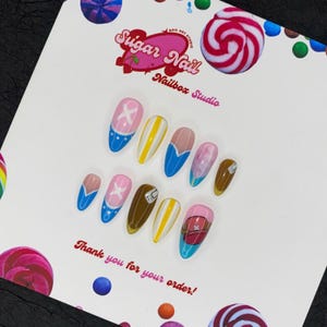 May include: A collection of custom nail art designs in a variety of colours and patterns. The designs include blue, pink, yellow, and brown nails with cartoon character art. The Sugar Nail Nailbox Studio logo and the text "Thank you for your order!" are visible.