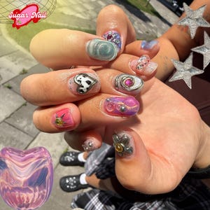 May include: Close-up of a hand with various nail art designs. The nails feature a range of colors, including pink, silver, and blue, with embellishments like hearts, gems, and floral accents. The text "Sugar Nail" is visible in the upper left corner.