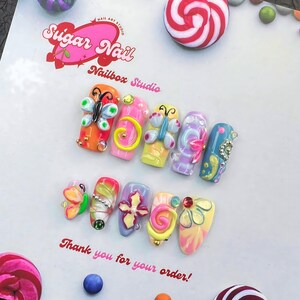 May include: A collection of colorful, decorated artificial nails. Each nail features unique designs, including butterflies, swirls, and floral accents. The nails are set against a white background with the text "Sugar Nail Nailbox Studio" and "Thank you for your order!"