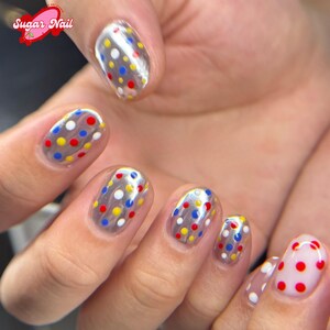 K&ouml;nnte beinhalten: Nahaufnahme von manik&uuml;rten N&auml;geln mit klarer Basis und bunten Tupfen in Rot, Gelb, Blau und Wei&szlig;. Die N&auml;gel haben ein gl&auml;nzendes Finish. Das Sugar Nail-Logo ist oben links sichtbar.