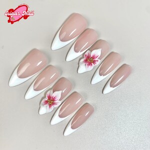 May include: A set of almond-shaped artificial nails with a white tip and a nude base. Some nails feature a 3D white flower with pink accents and gold beads. The image includes the text "Sugar Nail" in a pink logo.