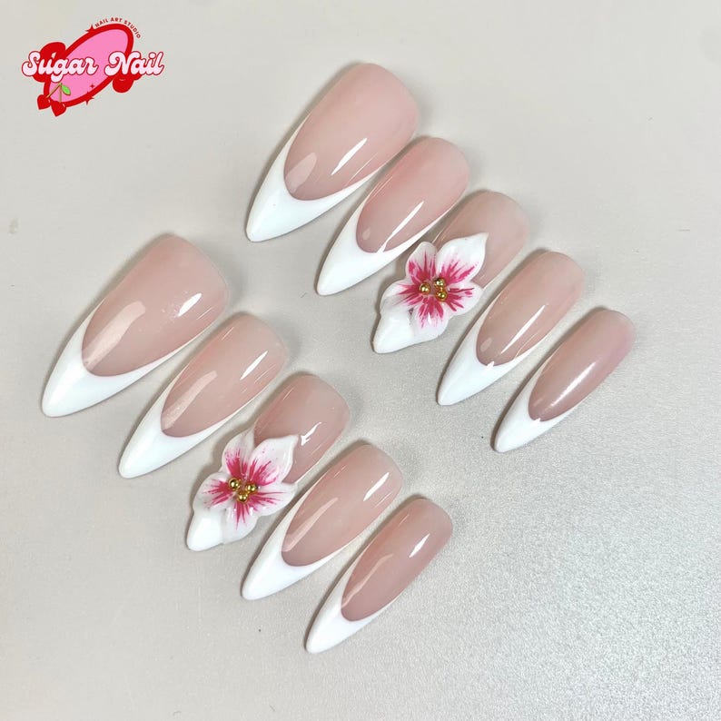 May include: A set of almond-shaped artificial nails with a white tip and a nude base. Some nails feature a 3D white flower with pink accents and gold beads. The image includes the text "Sugar Nail" in a pink logo.
