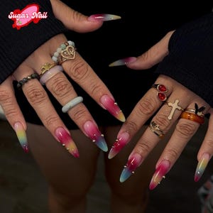 Puede incluir: Uñas largas en forma de estilete con un diseño degradado de rosa a amarillo, adornadas con tachuelas doradas. La imagen también muestra varios anillos en los dedos, incluyendo uno en forma de corazón y otro con una cruz. El texto "Sugar Nail" es visible.