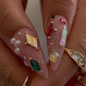May include: Close-up of long, almond-shaped nails painted a light pink. The nails are adorned with various colorful gemstones, including red, green, yellow, and iridescent crystals, and small gold studs. A gold ring with clear stones is also visible.