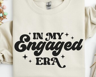 May include: A cream-colored sweatshirt with the phrase "IN MY Engaged ERA" in bold, black lettering. The text is stylized with a retro font and star accents. The sweatshirt is folded neatly on a white surface.