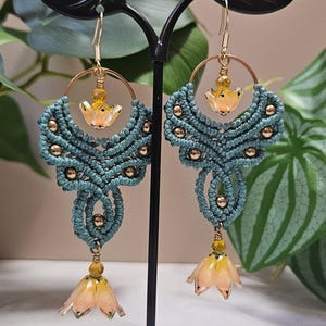 May include: Handmade macrame earrings in teal with gold accents. Each earring features a woven design with gold beads, a gold hoop, and a dangling flower-shaped charm in shades of yellow and peach. The earrings are displayed on a black stand.