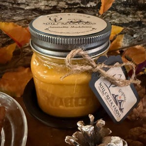 May include: A jar of Amaretto Madeleine scented candle with a silver lid and a twine bow. The jar has a black label with the words "Mill Creek Farms" and "Organic". The candle is on a black plate.