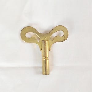 May include: A gold-toned metal clock winding key featuring a butterfly-shaped handle with two symmetrical cutouts and a cylindrical shaft with a single horizontal groove near the center, designed for manual winding mechanisms.