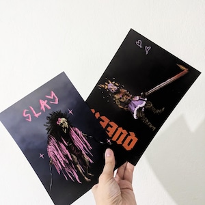 Puede incluir: Dos impresiones artísticas sostenidas por una mano. Una impresión presenta la palabra "SLAY" en rosa sobre una figura con un chal de plumas. La otra impresión tiene la palabra "QUEEN" en naranja, con una figura con corona y vestido púrpura.