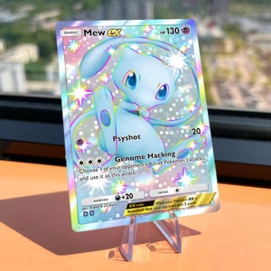 May include: A holographic Mew ex trading card featuring a light blue character illustration, 130 HP, and the attacks Psyshot and Genome Hacking. The card displays a rainbow foil finish with star patterns, set on a clear plastic display stand.