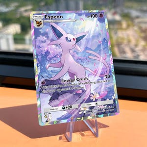 May include: A holographic Espeon Pokémon card, featuring a lavender-colored creature with large ears and a long tail. The card displays the text "Espeon", "Energy Crush", and "HP 100". The background shows a dreamy, floral landscape.