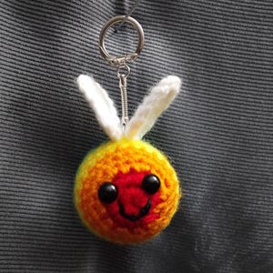 Puede incluir: Un llavero de ganchillo hecho a mano con una criatura redonda sonriente con una cara roja, un cuerpo naranja y amarillo y orejas de conejo blancas. El llavero tiene un anillo y una cadena de metal plateado.