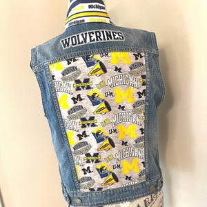 May include: A denim vest with a back panel featuring a repeating pattern of yellow and blue Michigan Wolverines logos and text. The word "WOLVERINES" is across the top. The vest is displayed on a mannequin.
