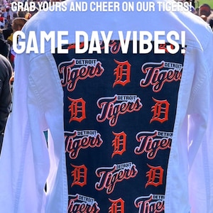 Könnte beinhalten: Weiße Jeansjacke mit einem marineblauen Rückenteil mit sich wiederholendem Detroit Tigers-Logo in Orange. Der Text "GAME DAY VIBES!" ist in Weiß dargestellt. Die Jacke wird in einem Stadion gezeigt.