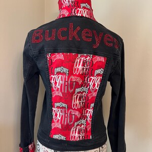 Könnte beinhalten: Schwarze Jeansjacke mit roten und weißen Stoffakzenten der Ohio State University. Das Wort "Buckeyes" ist mit roten Strasssteinen auf dem Rücken geschrieben. Die Jacke hat lange Ärmel und einen rot-weiß gemusterten Kragen und Manschetten.