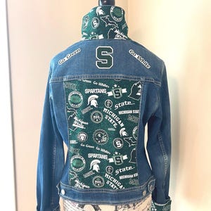 May include: A blue denim jacket with a green and white Michigan State Spartans design on the back. The jacket has a white "S" on the back and the words "Go Green" and "Go White" on the shoulders.