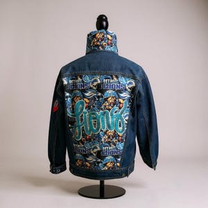 May include: A denim jacket with a Detroit Lions theme. The back features a vibrant design with the team's logo and the word "Lions" in a sparkling, turquoise script. The collar also has a matching pattern.