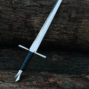 Op de afbeelding: Een tweesnijdend stalen zwaard met een lang, puntig lemmet, een rechte metalen pareerstang met gegraveerde details, een met zwart leer omwikkelde greep en een gepolijste metalen pommel die uitloopt in een hoekige punt aan de onderzijde.