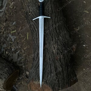 May include: A silver sword with a black handle and a crossguard, resting on a textured brown tree trunk. The sword has a pointed tip and a circular pommel. The image is taken from a high angle.