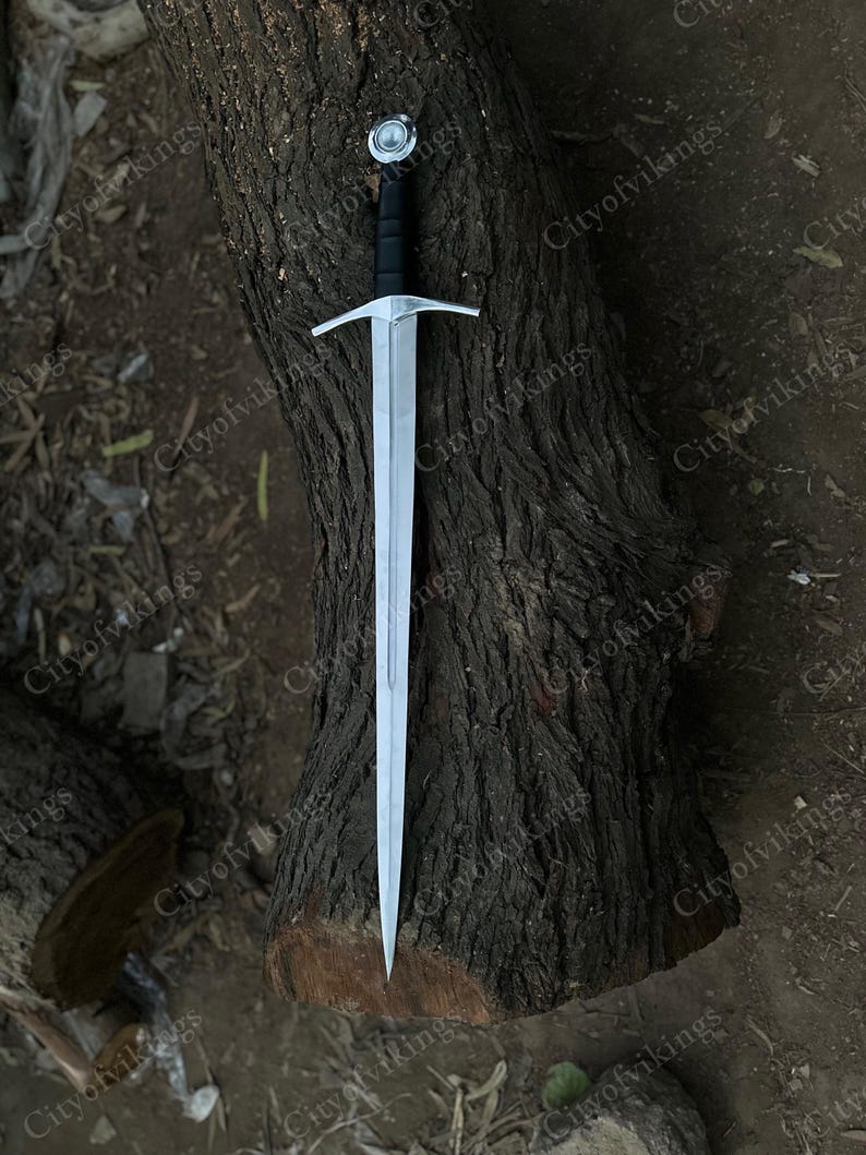 May include: A silver sword with a black handle and a crossguard, resting on a textured brown tree trunk. The sword has a pointed tip and a circular pommel. The image is taken from a high angle.