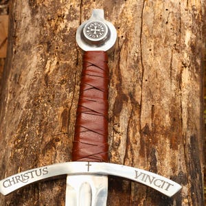 May include: A medieval sword with a silver blade and a brown leather-wrapped handle. The crossguard is inscribed with the words "CHRISTUS VINCIT". The pommel features a circular emblem with a cross design. The sword rests on a weathered wooden surface.