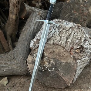 May include: A medieval sword with a silver blade and a black handle. The sword has a cross-shaped guard and a round pommel. The sword is resting on a large, weathered log. The text "CityofVikings" is visible in the background.
