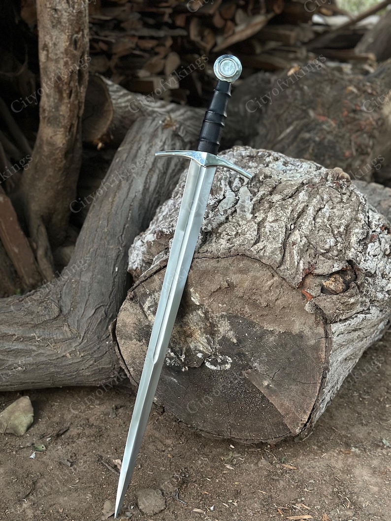 May include: A medieval sword with a silver blade and a black handle. The sword has a cross-shaped guard and a round pommel. The sword is resting on a large, weathered log. The text "CityofVikings" is visible in the background.