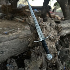 May include: A silver-colored sword with a long, pointed blade and a black handle. The sword features a crossguard and a circular pommel. The blade is resting on a weathered wooden surface, with the text "Cityofvikings" visible.