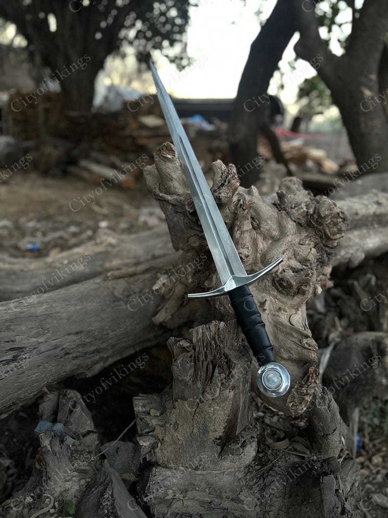 May include: A silver-colored sword with a long, pointed blade and a black handle. The sword features a crossguard and a circular pommel. The blade is resting on a weathered wooden surface, with the text "Cityofvikings" visible.