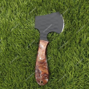 以下が含まれることがあります： テクスチャ加工されたヘッドと磨かれた木製のハンドルが付いた、小さな黒い斧。ハンドルは白のアクセントが付いた大理石模様です。斧は緑の草を背景に置かれています。