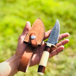 May include: A handcrafted hunting knife with a patterned steel blade, a light-colored handle, and a dark brown wooden base. The knife is accompanied by a brown leather sheath with a belt loop. The knife measures approximately 8 inches in total length.