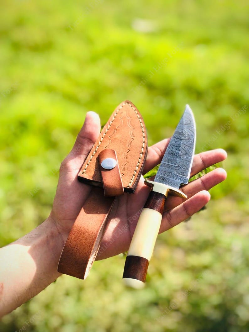 May include: A handcrafted hunting knife with a patterned steel blade, a light-colored handle, and a dark brown wooden base. The knife is accompanied by a brown leather sheath with a belt loop. The knife measures approximately 8 inches in total length.