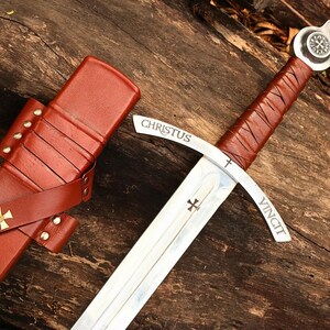 May include: A medieval sword with a brown leather-wrapped handle and a silver blade. The blade has the inscription "CHRISTUS VINCIT" and a cross symbol. The sword is next to a brown leather sheath with a gold cross.