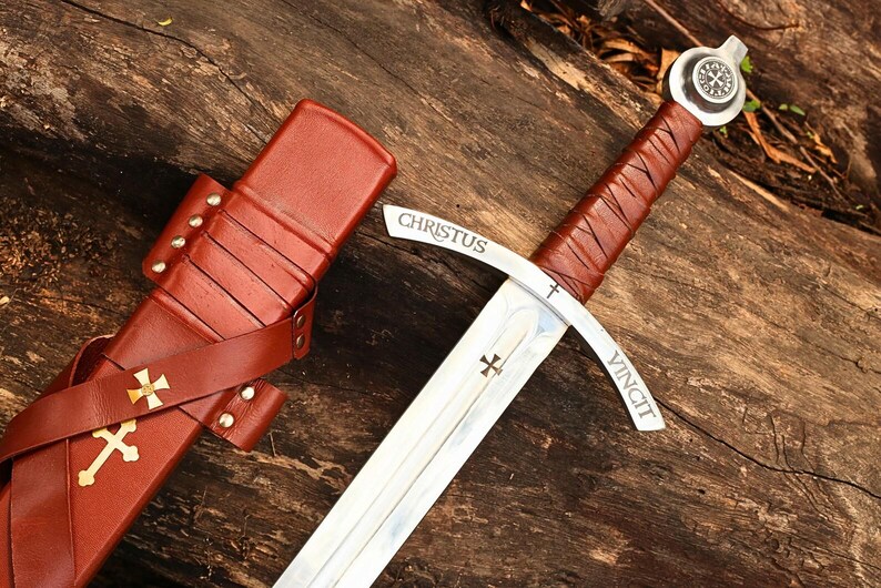 May include: A medieval sword with a brown leather-wrapped handle and a silver blade. The blade has the inscription "CHRISTUS VINCIT" and a cross symbol. The sword is next to a brown leather sheath with a gold cross.