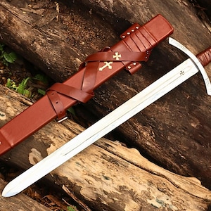 May include: A medieval-style sword with a silver blade and a brown leather-wrapped handle. The sword rests on a wooden surface next to a brown leather sheath adorned with gold crosses. The sheath is secured with straps and buckles.