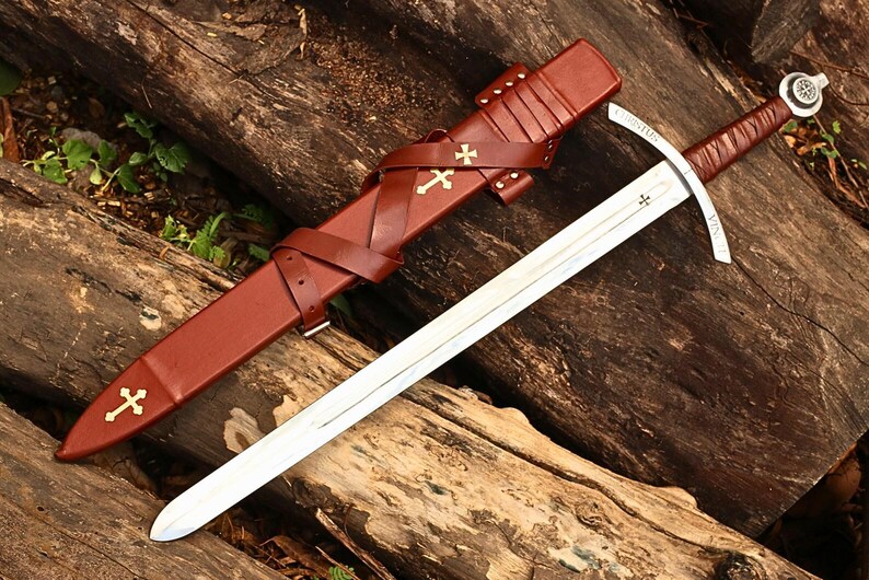 May include: A medieval-style sword with a silver blade and a brown leather-wrapped handle. The sword rests on a wooden surface next to a brown leather sheath adorned with gold crosses. The sheath is secured with straps and buckles.