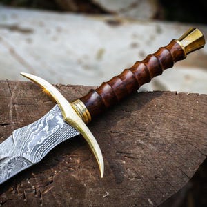 Peut inclure: Gros plan sur une épée avec une lame à motifs et un manche en bois. L'épée présente une garde et un pommeau dorés. Le manche est brun foncé et la lame présente un motif argenté et gris.