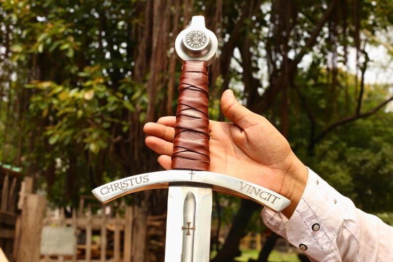 May include: A medieval-style sword with a silver blade and a brown leather-wrapped handle. The crossguard is inscribed with the words "CHRISTUS VINCIT". The sword features a cross emblem on the blade and a circular emblem on the pommel.