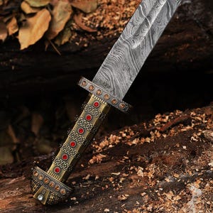 K&ouml;nnte beinhalten: Nahaufnahme eines dekorativen Dolches mit einer gemusterten Stahlklinge. Der Griff ist goldfarben und mit roten und klaren Edelsteinen verziert. Der Dolch liegt auf einem Bett aus Holzsp&auml;nen und einer dunklen Holzoberfl&auml;che.