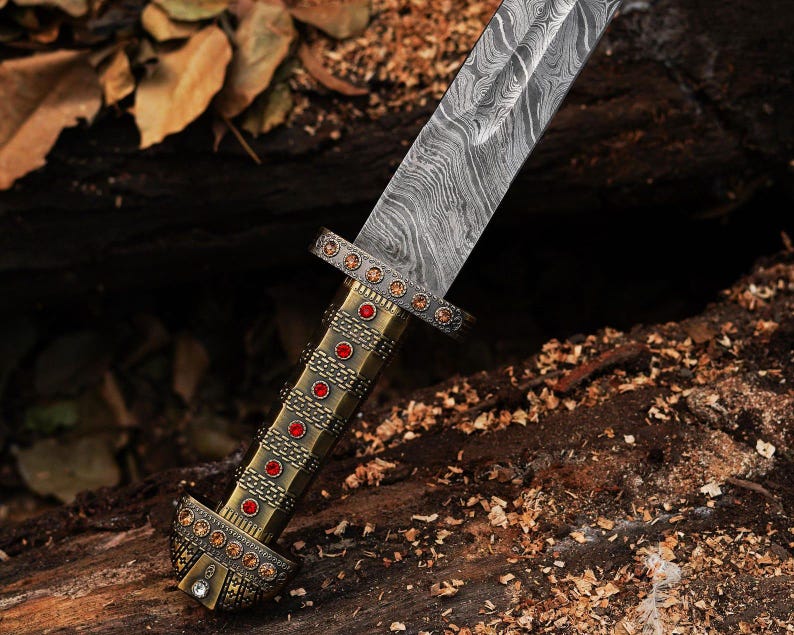 K&ouml;nnte beinhalten: Nahaufnahme eines dekorativen Dolches mit einer gemusterten Stahlklinge. Der Griff ist goldfarben und mit roten und klaren Edelsteinen verziert. Der Dolch liegt auf einem Bett aus Holzsp&auml;nen und einer dunklen Holzoberfl&auml;che.