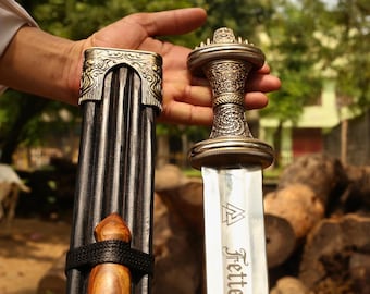 May include: A metal sword with an ornate, textured handle and a silver blade featuring a triangular symbol and the text Fetter. The sword is accompanied by a dark, vertically ribbed scabbard with decorative metal accents and a wooden detail.