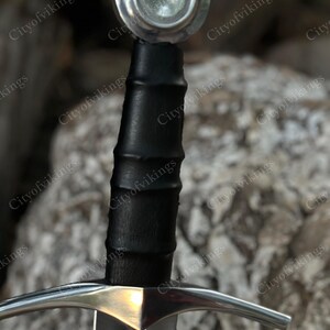 May include: Close-up of a medieval sword. The sword features a silver crossguard and a black leather-wrapped handle. The pommel is a silver disc. The blade is silver and the sword is set against a blurred background.