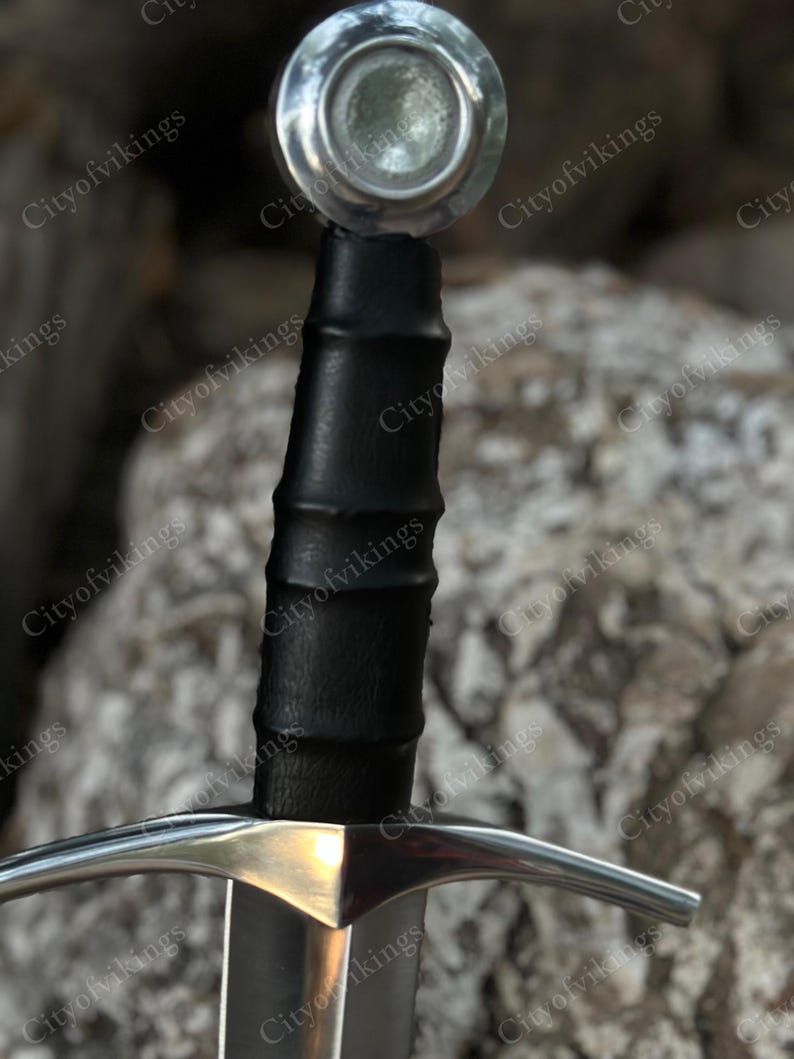 May include: Close-up of a medieval sword. The sword features a silver crossguard and a black leather-wrapped handle. The pommel is a silver disc. The blade is silver and the sword is set against a blurred background.